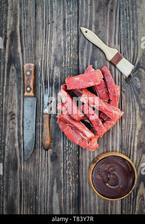 Matières premières côtes de boeuf désossé non cuit avec sauce bbq, pinceau, vintage et fourchette à viande couteau de boucher sur le dessus d'une table en bois rustique / arrière-plan. Capture d'image bof Banque D'Images