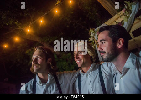 Trois jeunes hommes en armes autour de l'autre à une réception de mariage en plein air la nuit Banque D'Images