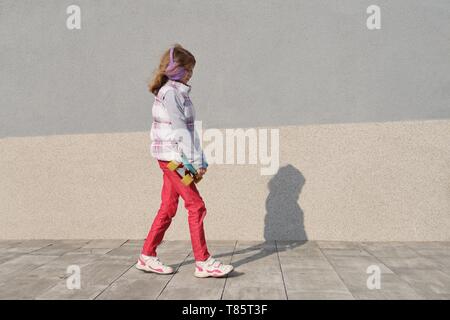 Petite fille monte un skateboard, dans la zone près de la maison, la saison du printemps, fond de mur gris. Banque D'Images