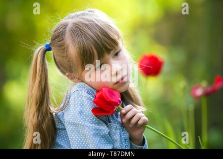 Cute pretty smiling enfant fille avec les yeux gris et les cheveux longs avec des fleurs tulipe rouge sur fond vert ensoleillé flou flou. L'amour à la nature d'hiver Banque D'Images