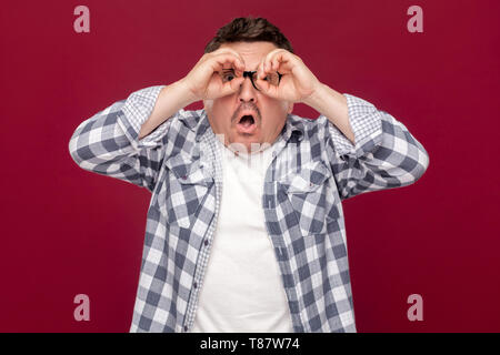 Portrait of business man in casual chemise à carreaux, les lunettes debout avec des jumelles sur les yeux et des gestes à visage choqué avec ind. Banque D'Images
