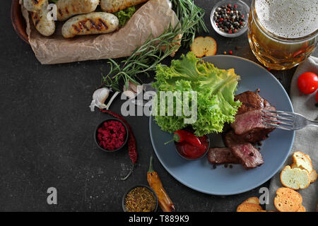 Chope de bière avec de délicieux steak grillé, des saucisses et des épices sur la table gris Banque D'Images