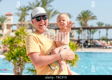 Heureux père tenant son fils joyeux en vacances, resort avec piscine et palmiers en arrière-plan, Portugal Banque D'Images