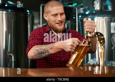 Vue avant de barmen masculins travaillant dans un pub et en injectant de l'ale dans le verre. Jeune homme barbu l'entretien de personnes dans la brasserie. Homme debout au comptoir, rire et parler. Concept de boisson. Banque D'Images