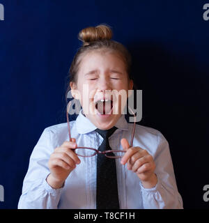 Fatigué d'écolière élémentaire de bâiller. Épuisé little girl holding lunettes. Enfant paresseux dans l'uniforme scolaire sur fond bleu foncé. Enfant endormi, leçon ennuyeuse, étudiant surmené Banque D'Images