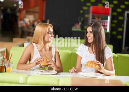 Vue de face de deux filles attirantes manger de délicieux hamburger et croissant dans le café. Brune et blonde sitting at table, regarder, parler et rire. Concept de la réunion. Banque D'Images