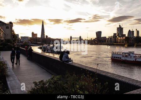 Les gens à pied et un couple s'asseoir et regarder le coucher du soleil derrière un grand nombre de bâtiments emblématiques de Londres et les ponts Banque D'Images