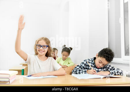 Les écoliers, les adolescents sitting at desk in classroom. Jolie, heureux lycéenne en bleu lunettes avec camera main posée. Garçon concentré l'écriture avec stylo dans cahier. Banque D'Images