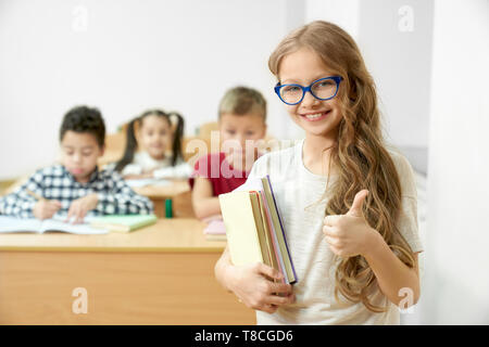 Gai, positif, cute schoolgirl standing in classroom, holding books et showing thumb up. Jolie enfant de l'école primaire dans les verres looking at camera, posant. Banque D'Images