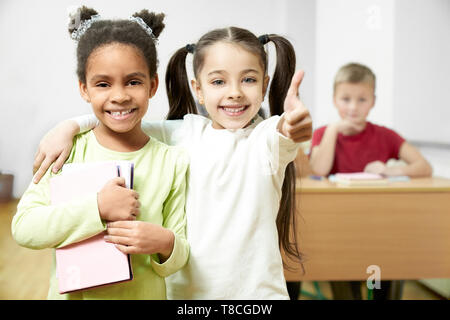 Jolies écolières standing in classroom, camarades showing Thumbs up. Cheerful, élèves positifs looking at camera, smiling, posant. Garçon assis au bureau sur l'arrière-plan. Banque D'Images