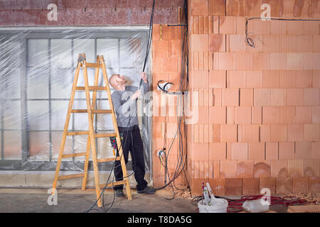 Électricien au travail l'installation de câbles dans le canal / groove avec boîtes électriques dans la maison inachevée construite de briques bloc d'argile. Pulli travailleur Banque D'Images