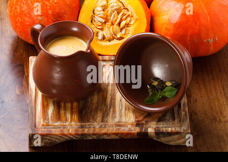 Soupe de potiron servie dans un pot/pot en argile, servie sur une planche de bois avec des citrouilles entières en arrière-plan. Banque D'Images