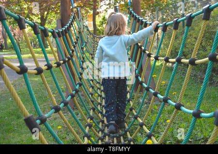 Cute little cca 3 ans, fille de marcher sur un pont de corde et net-comme la structure de jeu, sur une aire de printemps ensoleillée après-midi, vue de dos. Banque D'Images