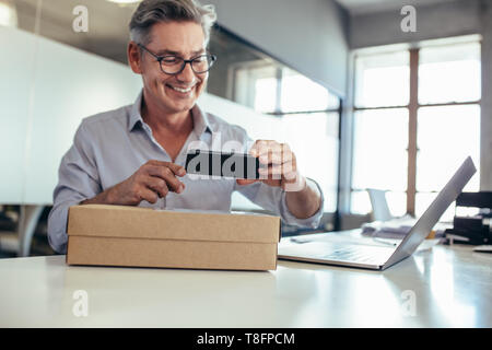 Smiling young man en tenant une boîte de livraison de numérisation sur son bureau. L'homme de prendre la photo d'un paquet. Banque D'Images