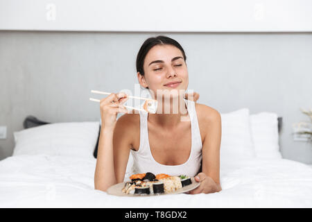 Belle jeune femme se détendre sur le lit chez lui, manger une assiette de sushi Banque D'Images