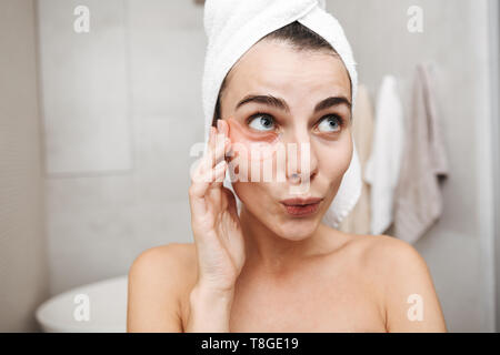 Gros plan d'une belle jeune femme avec une serviette sur la tête debout à la salle de bain, l'application de patchs oculaires Banque D'Images