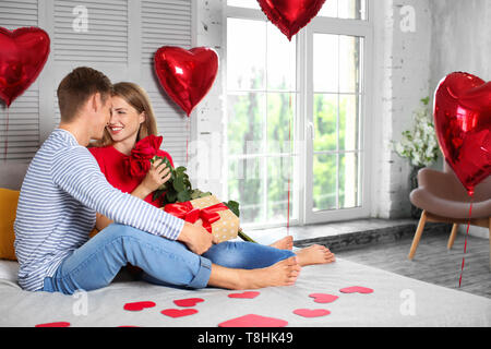 Jeune couple sympathique avec des roses rouges et gift box sitting on bed at home Banque D'Images
