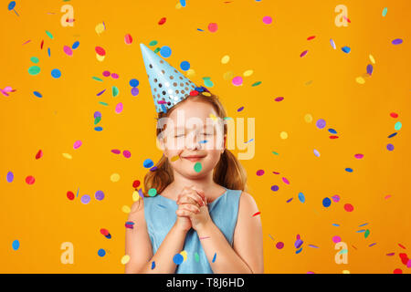 Cute little girl célèbre l'anniversaire. L'enfant ferma les yeux et fait un souhait dans la pluie de confettis. Closeup portrait sur fond jaune. Banque D'Images