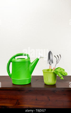Shot vertical d'un arrosoir vert avec un bec blanc à côté d'un pot vert avec une truelle, fourche de jardinage, et une paire de gants verts à l'intérieur. Sol en bois Banque D'Images