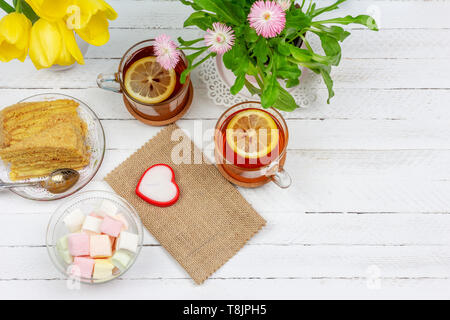 La vie toujours avec des tasses de thé, morceaux de gâteau, bouquet de fleurs roses dans un pot rose, guimauve et bougie en forme de cœur sur une table en bois blanc avec c Banque D'Images