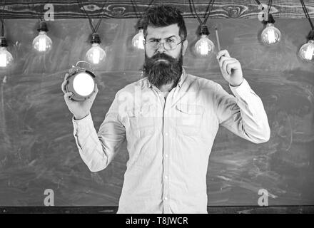 Enseignant dans les lunettes est titulaire d'un réveil et un stylo. Concept de discipline. Hipster barbu est titulaire d'réveil, tableau sur l'arrière-plan. Homme avec barbe et moustache sur la lettre, se tient dans la salle de classe. Banque D'Images