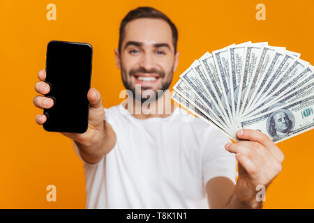 Photo de l'homme 30s dans des vêtements décontractés holding cell phone et le ventilateur d'argent sur fond jaune isolé Banque D'Images