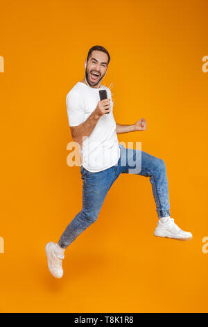 Photo de l'homme émotionnel 30s chanter pendant que l'aide d'écouteurs et le téléphone mobile plus isolé sur fond jaune Banque D'Images