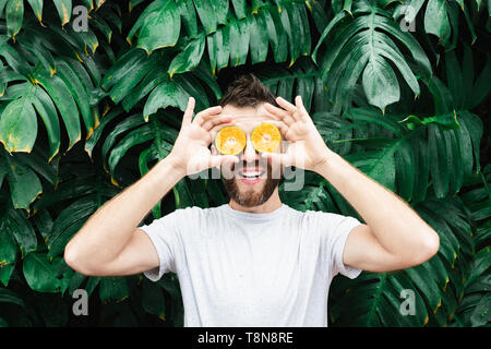 Jeune homme barbu tenant tranches de mandarine orange devant ses yeux, rire. Arrière-plan de feuilles tropicales, de copier l'espace. Banque D'Images