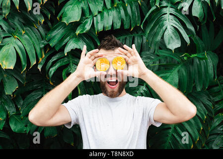 Jeune homme barbu tenant tranches de mandarine orange devant ses yeux, Laughing Out Loud. Arrière-plan de feuilles tropicales, de copier l'espace. Banque D'Images