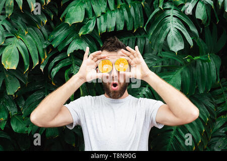 Jeune homme barbu tenant tranches de mandarine orange devant ses yeux, surpris sur son visage. Arrière-plan de feuilles tropicales, de copier l'espace. Banque D'Images