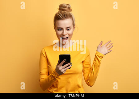 Le choc émotionnel des jeunes girl looking at mobile phone isolé sur fond jaune. la photo en gros. femme a lire informations intéressantes forment le gadget. Banque D'Images
