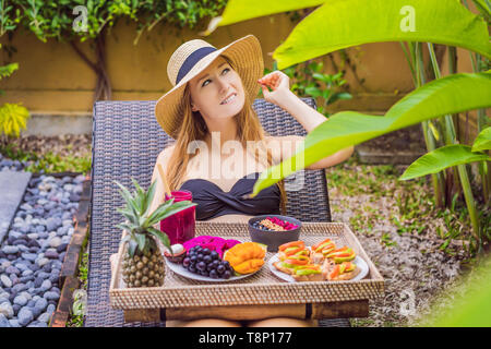 Young woman eating breakfast dans une chaise longue sur un plateau de fruits, petits pains, des sandwichs à l'avocat smoothie, bol de la piscine. Régime alimentaire sain d'été, végétalien Banque D'Images