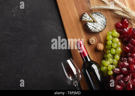 Bouteille de vin rouge, grappe de raisins, du fromage, des épis de blé et de verre sur planche de bois et fond noir. Vue de dessus avec l'exemplaire de l'espace. Banque D'Images