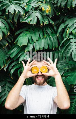 Jeune homme barbu tenant tranches de mandarine orange devant ses yeux, rire. Arrière-plan de feuilles tropicales, de copier l'espace. Banque D'Images