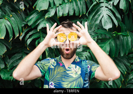 Jeune homme barbu tenant tranches de mandarine orange devant ses yeux, surpris sur son visage. Arrière-plan de feuilles tropicales, de copier l'espace. Banque D'Images