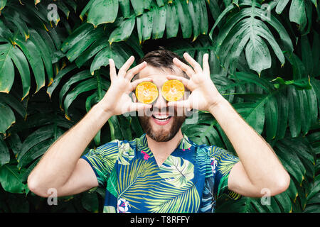 Jeune homme barbu tenant tranches de mandarine orange devant ses yeux, rire. Arrière-plan de feuilles tropicales, de copier l'espace. Banque D'Images