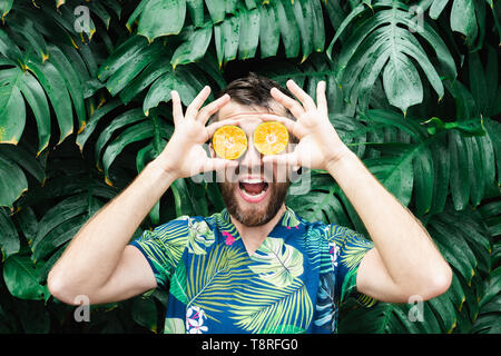 Jeune homme barbu tenant tranches de mandarine orange devant ses yeux, Laughing Out Loud. Arrière-plan de feuilles tropicales, de copier l'espace. Banque D'Images