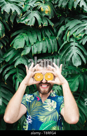 Jeune homme barbu tenant tranches de mandarine orange devant ses yeux, rire. Arrière-plan de feuilles tropicales, de copier l'espace. Banque D'Images