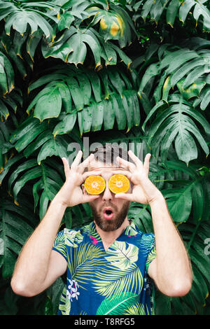 Jeune homme barbu tenant tranches de mandarine orange devant ses yeux, surpris sur son visage. Arrière-plan de feuilles tropicales, de copie, de l'espace v Banque D'Images