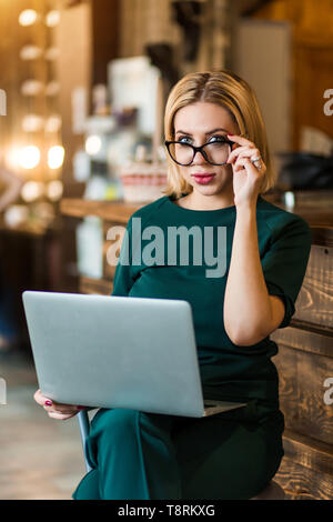 Assez sérieuse femme élégante avec de courts cheveux blonds, vert émeraude, le port de boucles d'adapter et de verres de couleur sur la plage près de réception dans Banque D'Images