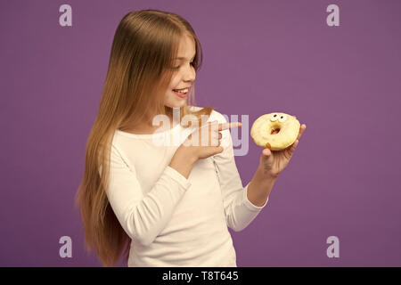 Happy kid pointer du doigt donut sur fond violet. Petite fille avec bague donut glacé sur fond violet. La mauvaise alimentation. Sourire de l'enfant avec la malbouffe. La nourriture et le dessert. L'enfance et de l'enfants. Banque D'Images