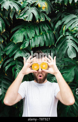 Jeune homme barbu tenant tranches de mandarine orange devant ses yeux, surpris sur son visage. Arrière-plan de feuilles tropicales, de copie, de l'espace v Banque D'Images