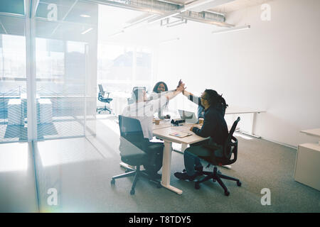 Groupe diversifié de collègues de travail de rire et de plafonnement élevé, tandis qu'assis autour d'une table à l'intérieur d'un bureau des murs de verre, Banque D'Images