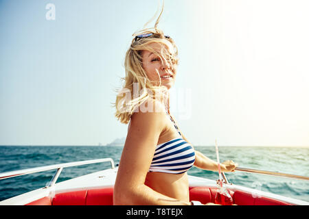 Smiling young woman in a bikini top détente sur un bateau avec un verre sur une journée ensoleillée pendant ses vacances d'été Banque D'Images