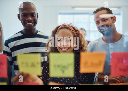 Groupe de designers Smiling divers lecture notes adhésives sur un mur de verre dans un bureau moderne pendant une session de remue-méninges Banque D'Images