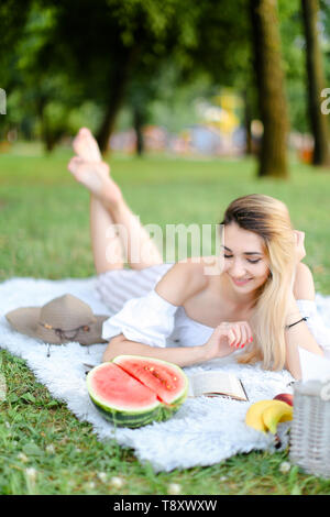 Jeune blonde woman lying on sac en park, reading book près de pastèque et hat. Concept de loisirs, vacances d'été et pique-nique. Banque D'Images