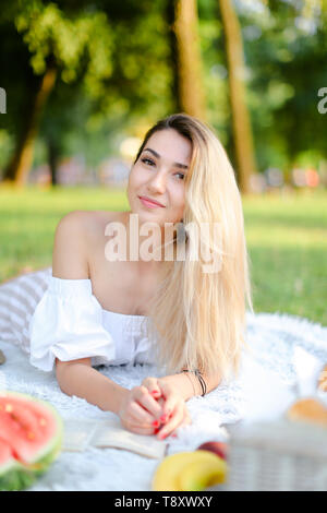 Jeune femme couchée sur plaid près de pastèque dans le parc et la lecture de livre. Concept de pique-nique sur la nature, les vacances et les loisirs. Banque D'Images