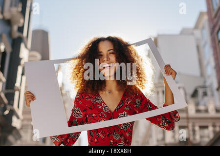 Belle robe woman looking at camera avec cadre photo vide. Jolie femme avec photo frame standing en plein air dans la ville. Banque D'Images