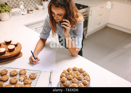 Vue de dessus de la pâtisserie de prendre des commandes par téléphone dans la cuisine. Femme chef de l'ordre d'écriture tout en parlant sur téléphone mobile. Banque D'Images