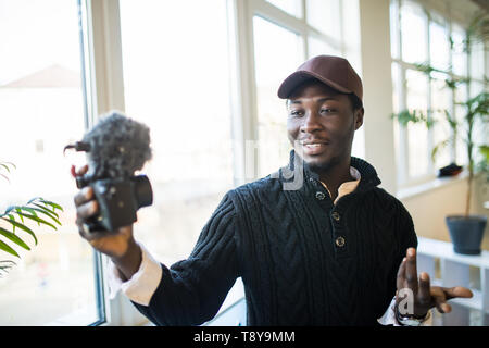 Jeune homme africain blogueur vidéo crée du contenu vidéo pour son canal. Heureux guy shoots video streaming pour les utilisateurs tout en restant assis dans le bureau. Banque D'Images